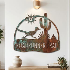 Peut inclure: Une enseigne murale en métal semi-circulaire avec une finition patinée rouille brune et bleu sarcelle oxydée. Le motif représente un grand géocoucou, un cactus saguaro et un soleil. Le texte ROADRUNNER TRAIL est découpé dans le métal.
