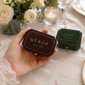 May include: A dark brown velvet ring box with gold foil lettering E & J and the date 05.16.2026, featuring a decorative border. Beside it sits a dark green velvet ring box with gold lettering A L and the date 05.20.2026, both with gold clasps.