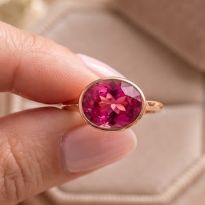 May include: A gold ring featuring a large, oval-cut, vibrant pink gemstone set in a smooth bezel setting. The band is thin and polished, showcasing the faceted surface of the stone which reflects light, creating a bright and clear appearance.