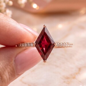 May include: A rose gold ring featuring a kite-shaped deep red gemstone as the centre stone. The thin band is adorned with small, clear pavé-set diamonds along the sides, creating a delicate and refined appearance for this piece of jewellery.