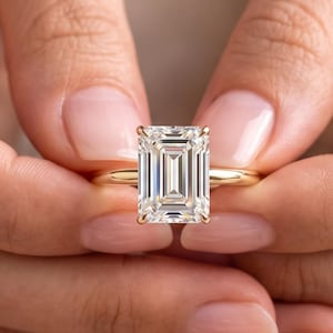 May include: A gold ring featuring a large, rectangular emerald-cut clear gemstone set in a four-prong setting. The band is a simple, polished gold metal. The stone displays clear facets and reflects light, centred on a thin, smooth gold band.