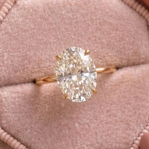 May include: A gold ring featuring a large, oval-cut, clear faceted gemstone set in a four-prong setting. The ring is displayed inside an octagonal, dusty rose-colored velvet jewelry box with a central slot for the band.