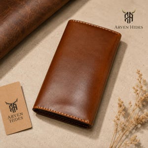 May include: A rectangular brown leather wallet with visible stitching along the edges. The wallet features a smooth, polished finish. A small tan tag is present with the text Arven Hides and a bull logo, which also appears in the upper right corner.