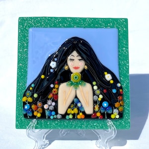 Op de afbeelding: Een groene ingelijste glazen tegel met een vrouw met lang zwart haar die een boeket gekleurde bloemen vasthoudt. De vrouw glimlacht en heeft een lichtblauwe achtergrond.