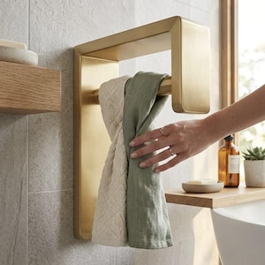 Könnte beinhalten: Ein wandmontierter Handtuchhalter aus gebürstetem Messing mit rechteckigem Rahmen und einer horizontalen Stange. Zwei gefaltete Handtücher hängen über der Stange, eines in strukturiertem Creme und das andere in einfarbigem Salbeigrün.