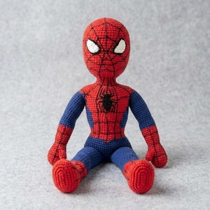 May include: A handmade crochet doll of a superhero character featuring a red and blue color scheme. The doll has a red head and torso with a black web pattern, white eyes, and blue arms and legs. A small black spider emblem is centered on the chest.