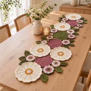May include: A long, decorative table runner crafted from crocheted floral motifs in cream, dusty rose, and olive green. The runner features large, scalloped-edge cream flowers, solid circular dusty rose discs, and small green crocheted leaves.