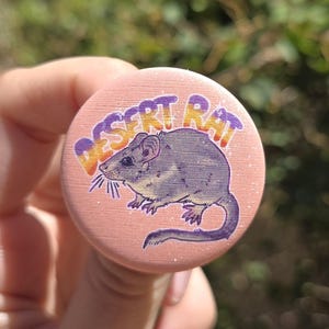 May include: A circular pin-back button with a light peach background featuring a central illustration of a grey rat. Above the rat, the text DESERT RAT is printed in a gradient of purple, orange, and yellow. The surface has a subtle glitter finish.