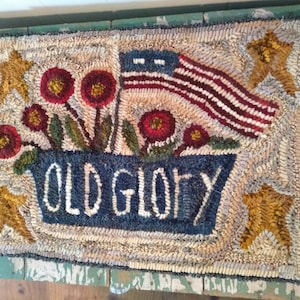 Könnte beinhalten: Ein handgefertigter Teppich mit einer blau-weißen amerikanischen Flagge, roten Blumen in einem blauen Topf und den Worten "Old Glory" in weißen Buchstaben.