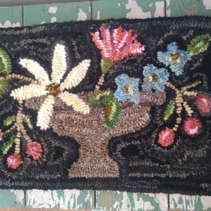 May include: A rectangular, hand-hooked rug with a black background. The rug features a central vase filled with flowers in shades of pink, blue, and white. Green leaves and red berries are also present, creating a floral design.
