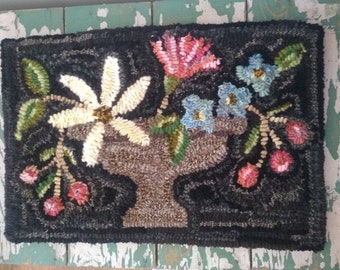 Puede incluir: Una alfombra rectangular, hecha a mano con un fondo negro. La alfombra presenta un jarrón central lleno de flores en tonos rosa, azul y blanco. También hay hojas verdes y bayas rojas, creando un diseño floral.
