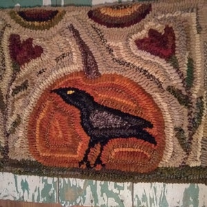 Könnte beinhalten: Ein schwarzer Rabe steht vor einem großen orangefarbenen Kürbis mit einem braunen Stiel. Der Kürbis ist von grünen Blättern und braunen Ranken umgeben. Der Hintergrund ist ein beige-brauner Teppich mit einem Blumenmuster.