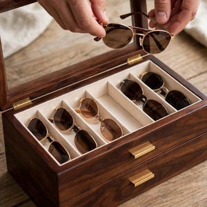 May include: A dark wood storage box with a glass lid and a pull-out drawer, featuring multiple interior compartments lined with cream-coloured fabric. The box holds several pairs of sunglasses with gold-toned frames and brown tinted lenses.