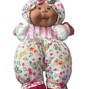 May include: A doll with a round face, brown eyes, and yarn hair, wearing a white bonnet with a pink cherry and floral print. The doll is dressed in a matching white romper featuring pink stripes, cherries, and flowers, with pink fabric shoes.