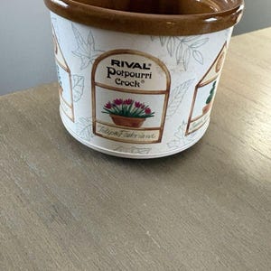 May include: A round ceramic Rival Potpourri Crock with a brown glazed rim and a white body. The exterior features a repeating graphic design of arched windows containing potted plants and the text Tulipa Fosteriana.