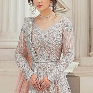 May include: A peach-colored long-sleeved gown featuring intricate silver embroidery and beadwork throughout the bodice and sleeves. The garment includes a matching sheer dupatta draped over one shoulder and a thin silver belt at the waist.