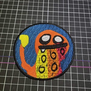 May include: A circular embroidered patch featuring an orange creature with a yellow tail tip, white eyes, and a black mouth. The creature has a multi-colored body with yellow, orange, and pink sections, set against a blue background with a wave pattern.