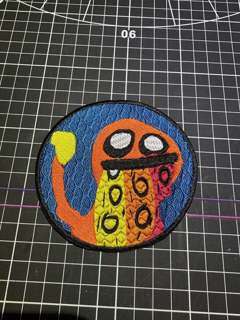 May include: A circular embroidered patch featuring an orange creature with a yellow tail tip, white eyes, and a black mouth. The creature has a multi-coloured body with yellow, orange, and pink sections, set against a blue background with a wave pattern.