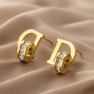Peut inclure: Une paire de boucles d'oreilles à tige dorées en forme de lettre D stylisée. Chaque boucle d'oreille intègre un élément circulaire incrusté de petits cristaux clairs et étincelants au centre du motif de la lettre, avec une tige de fixation.