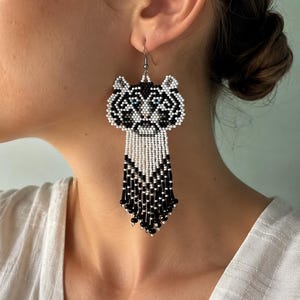 May include: A beaded dangle earring featuring a stylized tiger head design in black, white, and grey beads with small blue accents for the eyes. The earring includes a long, multi-strand fringe made of white and black beads hanging from the base.