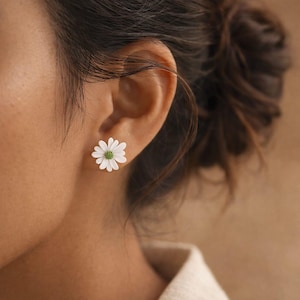 Peut inclure: Une boucle d'oreille à tige en forme de fleur de marguerite avec des pétales blancs et un centre vert texturé. La boucle d'oreille est placée sur un lobe d'oreille, mettant en valeur le motif floral avec une vue claire des pétales blancs.