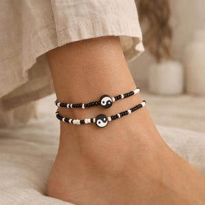 Op de afbeelding: Twee enkelbandjes met kleine zwarte en witte kraaltjes in een herhalend patroon. Elk enkelbandje is in het midden voorzien van een ronde, platte bedel met het zwart-witte yin yang symbool. De sieraden worden rond de enkel gedragen.