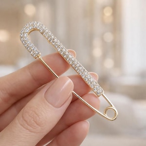Peut inclure: Une broche épingle à nourrice dorée dotée d'une longue tige droite incrustée de nombreuses petites pierres transparentes et scintillantes. L'épingle en métal présente une finition polie, un ressort classique à la base et une pointe acérée.