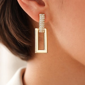 Peut inclure: Une boucle d'oreille pendante dorée composée d'un pendentif rectangulaire suspendu à une barre verticale. La barre supérieure est incrustée de petites pierres claires et scintillantes, tandis que le cadre rectangulaire inférieur est lisse.