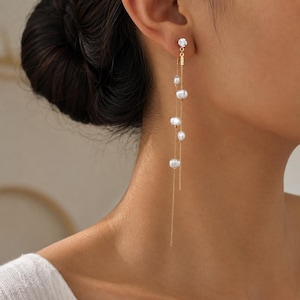 May include: A long gold-tone chain earring featuring a small clear stud at the top and multiple white pearls suspended at varying lengths along two delicate dangling chains that hang down from the earlobe in a layered, vertical arrangement.