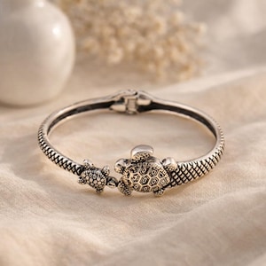 Könnte beinhalten: Ein silberfarbener Klapparmreif mit zwei Meeresschildkröten-Motiven und detaillierten Panzerstrukturen. Das Armband weist an den Seiten ein strukturiertes Rautenmuster auf und verfügt über einen sicheren Verschluss mit oxidiertem Finish.