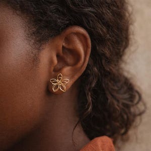 Peut inclure: Une boucle d'oreille à tige dorée en forme de fleur à cinq pétales, fabriquée à partir de fines boucles de fil continu formant un design ouvert et aéré. La boucle d'oreille est placée sur le lobe, révélant une silhouette florale métallique.