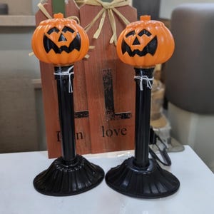 May include: Two identical Halloween decorations featuring orange plastic jack-o'-lantern heads with black painted facial features, mounted on tall black cylindrical posts with fluted circular bases, each secured with a thin white string.