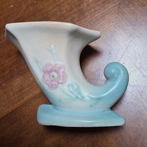 May include: A ceramic cornucopia-shaped vase featuring a matte cream-colored body with a soft blue base and a curled tail. The front of the vessel is decorated with a hand-painted pink flower with a yellow center and light green leaves.