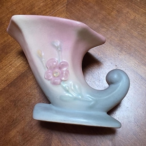 May include: A ceramic cornucopia-shaped vase featuring a soft gradient of pastel pink at the top and light blue at the base. The exterior is decorated with a raised, molded floral design in pink and yellow, accented by green leaves.