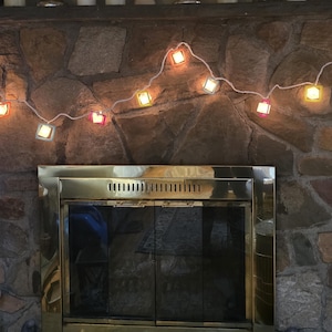 May include: A string of square, colorful lights hangs against a stone fireplace. The lights are encased in clear, square frames with a variety of colors, including yellow, orange, and pink. The fireplace has a brass-colored metal frame around the glass doors.