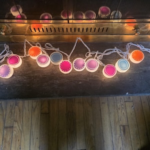 May include: A string of decorative lights with colorful, round shades. The shades are in various colors, including pink, orange, blue, and purple. The lights are strung on a white wire and are placed on a dark surface.