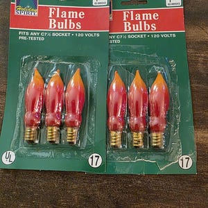 May include: Two packages of flame-shaped light bulbs. Each package contains three bulbs with a red and orange flame design. The packaging is green and red, with the text "Flame Bulbs" and "Holiday Spirit".