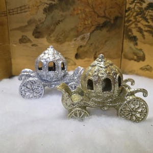 May include: Two decorative carriage ornaments, one silver and one gold, sit on a white surface. The carriages have intricate wheel and body details, with arched windows. The background features a gold-toned, Asian-inspired screen.