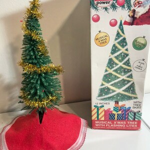 May include: A 12-inch musical Christmas tree with flashing lights and a red tree skirt. The tree is decorated with gold tinsel and plays three Christmas carols. The box reads "Model Power Musical X'mas Tree with Flashing Lites".