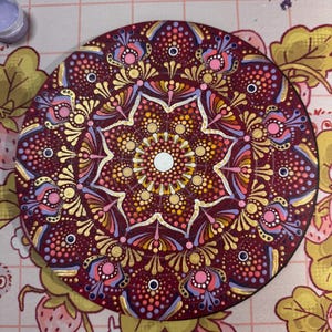 May include: A circular wooden plaque featuring a hand-painted mandala design in deep burgundy, gold, light pink, and lavender. The intricate pattern consists of concentric circles of dots and petal-like shapes radiating from a central white dot.