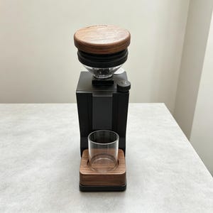May include: A black coffee grinder featuring a dark wood lid on the hopper and a matching dark wood base. The device includes a clear glass catch cup positioned on the wooden base, a black adjustment dial on the top, and a sleek, vertical body.