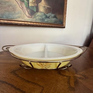 Könnte beinhalten: Eine ovale, unterteilte Servierschale aus Milchglas mit hellgelber Außenseite und einem Weizenährenmuster. Die Schale befindet sich in einem polierten Metallgestell, das mit zwei seitlichen Griffen und vier geschwungenen Füßen ausgestattet ist.