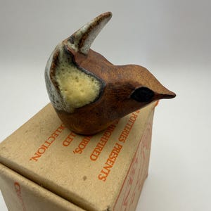 Può includere: Una piccola statuetta di uccello in ceramica con corpo marrone, una macchia gialla sull'ala e coda bianca, appoggiata su una scatola di cartone. La scatola presenta un testo arancione che recita POTTERIES PRESENTS, HIGHFIRED, BIRDS, HANDCRAFTED e COLLECTION.