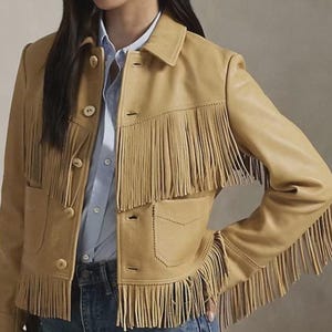 May include: A tan leather jacket featuring long fringe detailing across the chest and sleeves. The jacket includes a classic collar, a front button closure with light-coloured buttons, and two front pockets with stitched edges and a pointed flap design.