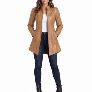 May include: A tan leather jacket with a mandarin collar, a full-length front zipper, and vertical seam detailing. The jacket is worn over a white top and dark blue denim jeans, paired with black leather ankle boots with block heels.