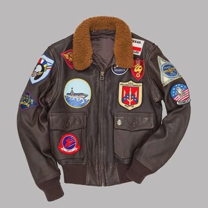 Pode incluir: Uma jaqueta de voo de couro marrom escuro com gola de pele sintética marrom, zíper frontal e dois bolsos com aba. A jaqueta é adornada com vários emblemas bordados coloridos em estilo militar, incluindo textos como Seabees, Texas e Tomcat.