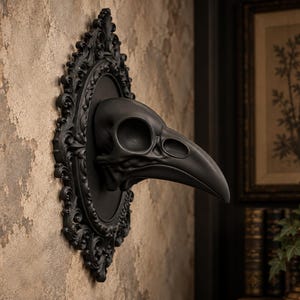 Puede incluir: Una escultura de cráneo de ave en negro mate montada sobre un marco ovalado a juego con intrincados detalles de volutas. El cráneo presenta un pico curvo prominente y cuencas oculares huecas, extendiéndose hacia afuera desde el centro de la placa.