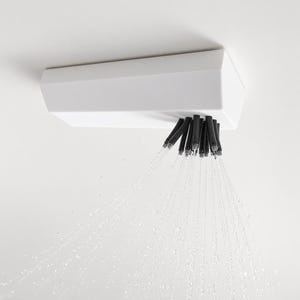 Pode incluir: Um chuveiro retangular branco montado numa superfície, com um conjunto de bicos pretos que emitem múltiplos jatos de água que fluem para baixo num padrão diagonal contra um fundo branco liso.