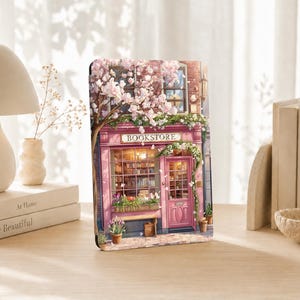May include: A rectangular decorative plaque featuring a detailed illustration of a pink bookstore storefront with a large tree covered in light pink blossoms, a glass window displaying shelves of books, and the word BOOKSTORE printed above the door.