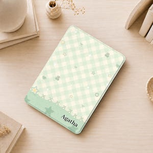 May include: A rectangular tablet case featuring a light green and white gingham pattern decorated with small stars, bows, and circular icons. The bottom section has a solid green band with a decorative stitched border and the name Agatha printed.